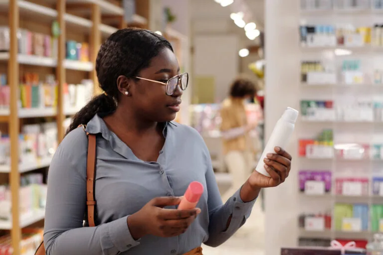 Young African American woman in eyeglasses holding two spray cans containing deodorant while choosing beauty products